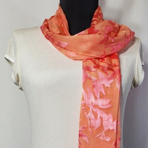 May include: A peach-colored scarf with a floral pattern in a darker shade of pink. The scarf is made of a sheer fabric and has a silky feel.