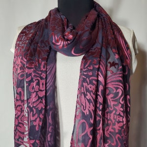 May include: A sheer scarf with a dark purple base and a vibrant magenta floral pattern. The scarf is draped over a mannequin, showcasing its flowing design and intricate details. This accessory adds a touch of elegance.