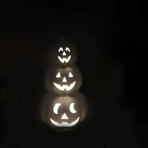 May include: A stack of three white ceramic jack-o'-lanterns with carved faces, illuminated from within. The pumpkins are stacked on top of each other, creating a festive Halloween decoration. The background is dark.
