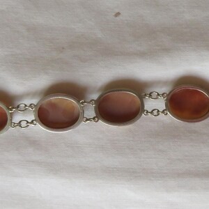 May include: A silver-toned bracelet featuring oval and rectangular reddish-brown stones. The stones are set in individual silver frames and connected by small silver chain links. The bracelet is displayed on a white fabric surface.
