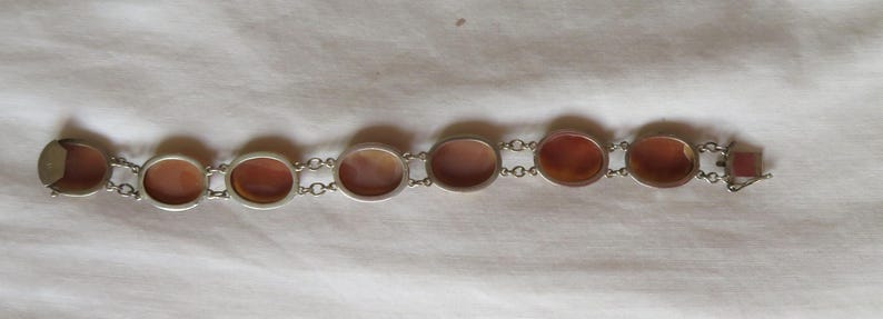May include: A silver-toned bracelet featuring oval and rectangular reddish-brown stones. The stones are set in individual silver frames and connected by small silver chain links. The bracelet is displayed on a white fabric surface.