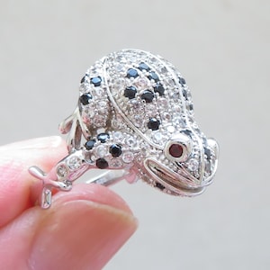 Op de afbeelding: Een zilverkleurige ring in de vorm van een kikker. Het lichaam van de kikker is bedekt met kleine, heldere en zwarte stenen, met rode stenen voor de ogen. De ring wordt ondersteund door de poten van de kikker.