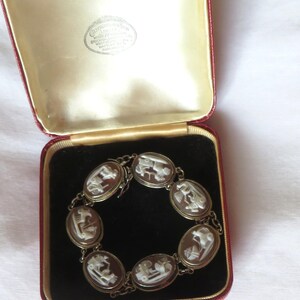 May include: An antique cameo bracelet displayed in a red velvet lined box. The bracelet features oval cameos with white relief figures set in a gold-toned metal chain. The box has a cream-colored satin lining.