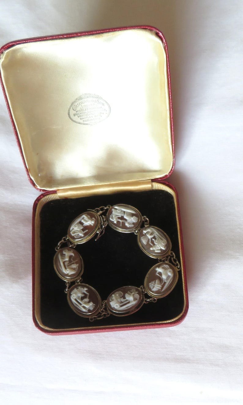 May include: An antique cameo bracelet displayed in a red velvet lined box. The bracelet features oval cameos with white relief figures set in a gold-toned metal chain. The box has a cream-colored satin lining.