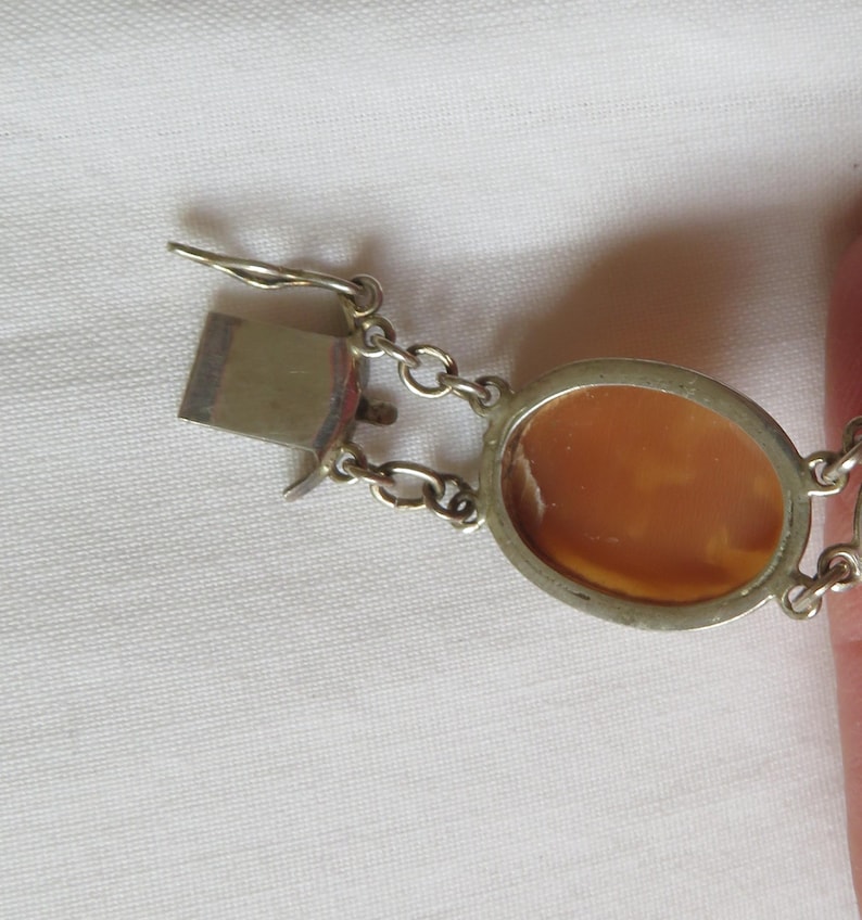 May include: A silver-toned bracelet with oval cameo pendants. The bracelet features a clasp and chain links. The cameo pendants have a light brown color and are set in a silver-toned metal frame. The bracelet is on a white surface.