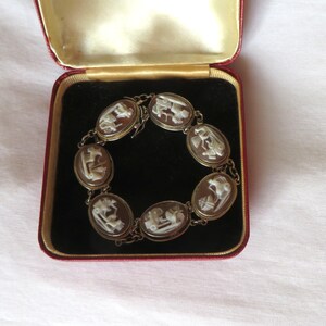 May include: A vintage cameo bracelet displayed in a red velvet jewelry box. The bracelet features oval-shaped cameos with white relief carvings set in a gold-toned metal. The box has a gold interior and a red exterior.