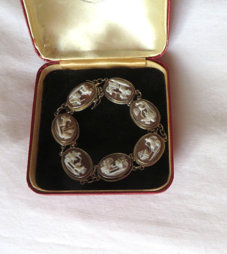 May include: A vintage cameo bracelet displayed in a red velvet jewelry box. The bracelet features oval-shaped cameos with white relief carvings set in a gold-toned metal. The box has a gold interior and a red exterior.