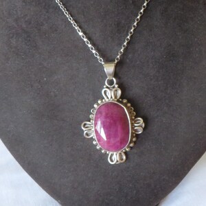 May include: A silver necklace featuring a large, oval, ruby-coloured pendant. The pendant is set in a decorative silver frame with ornate detailing. The necklace is displayed on a dark grey surface.