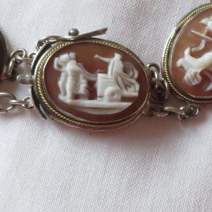 May include: A silver-toned bracelet featuring oval cameo pendants. Each pendant showcases a detailed white relief against a reddish-brown background, depicting classical figures. The bracelet is connected by small chain links.