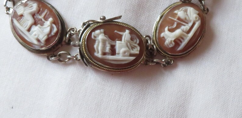 May include: A silver-toned bracelet featuring oval cameo pendants. Each pendant showcases a detailed white relief against a reddish-brown background, depicting classical figures. The bracelet is connected by small chain links.