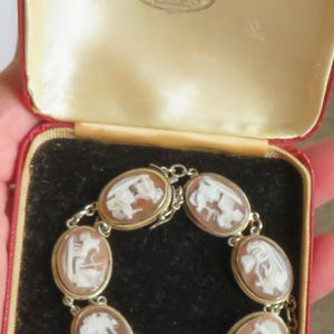 May include: A vintage cameo bracelet with seven oval-shaped cameos set in gold-tone metal. Each cameo features a white relief carving against a brown background. The bracelet is displayed in a red and cream jewelry box.