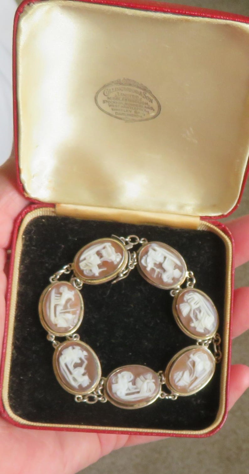 May include: A vintage cameo bracelet with seven oval-shaped cameos set in gold-tone metal. Each cameo features a white relief carving against a brown background. The bracelet is displayed in a red and cream jewelry box.