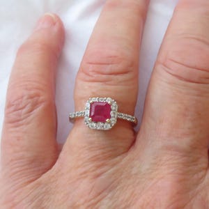 以下が含まれることがあります： 正方形の深い赤色の宝石を、より小さな透明な宝石のハローで囲んだ、金色のリング。 リングは、さらに小さな宝石が付いたバンドにセットされています。 リングは指にあります。