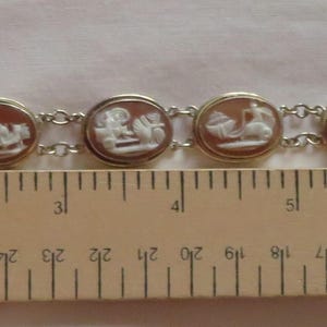 May include: A vintage bracelet featuring seven oval cameos with white relief figures set in gold-toned frames, connected by silver-toned links. The bracelet is approximately 7 inches long.