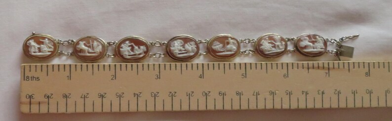 May include: A vintage bracelet featuring seven oval cameos with white relief figures set in gold-toned frames, connected by silver-toned links. The bracelet is approximately 7 inches long.