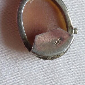 May include: Close-up of a silver pendant with a cameo design. The oval-shaped pendant features a light brown cameo set within a silver frame. The back of the pendant has the number "925" stamped on it. The pendant is on a white surface.