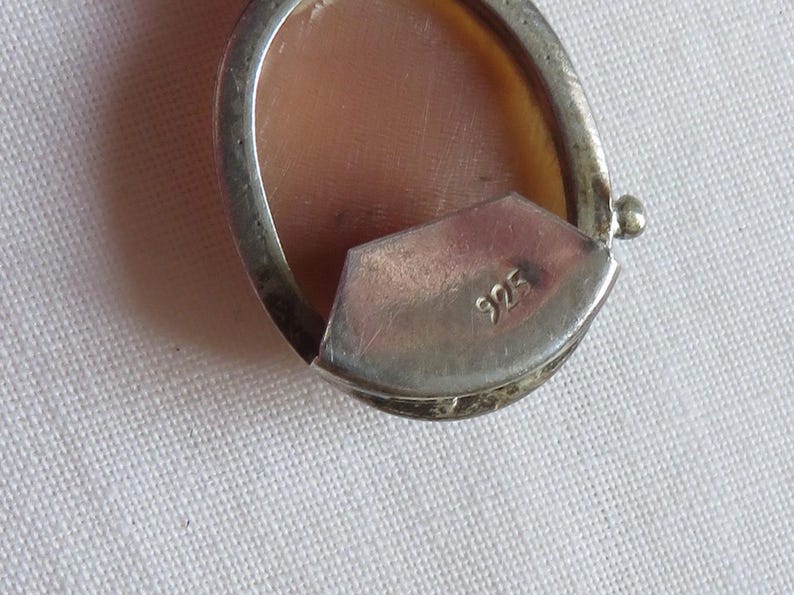 May include: Close-up of a silver pendant with a cameo design. The oval-shaped pendant features a light brown cameo set within a silver frame. The back of the pendant has the number "925" stamped on it. The pendant is on a white surface.
