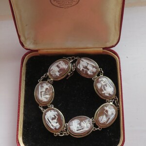 May include: An antique bracelet with oval cameo stones, each featuring a white relief carving against a brown background. The bracelet is set in a silver-toned metal and is displayed in a red and tan jewelry box. The box has text from Collingwood & Son.