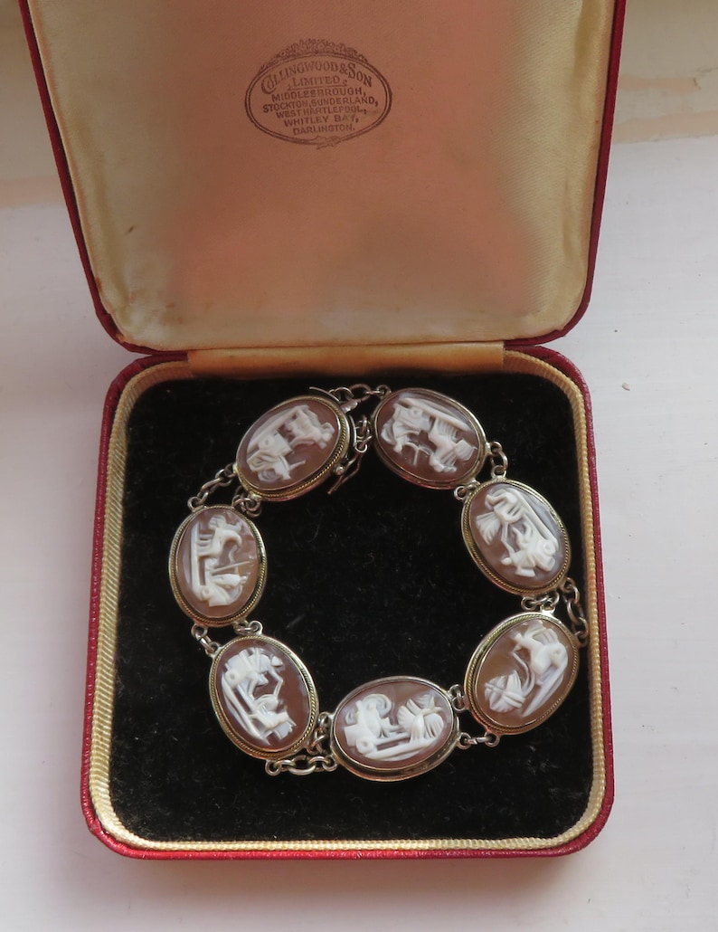 May include: An antique bracelet with oval cameo stones, each featuring a white relief carving against a brown background. The bracelet is set in a silver-toned metal and is displayed in a red and tan jewelry box. The box has text from Collingwood & Son.