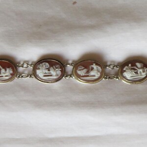 May include: A vintage bracelet featuring oval cameos with white relief figures set in gold-toned frames. The cameos are connected by a delicate silver chain, and the bracelet has a clasp closure. The bracelet is displayed on a white surface.