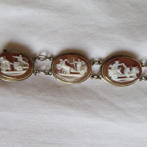 May include: A vintage bracelet featuring oval cameos with detailed white relief carvings set in gold-toned frames. The cameos depict classical figures and scenes, connected by delicate silver-toned chain links.