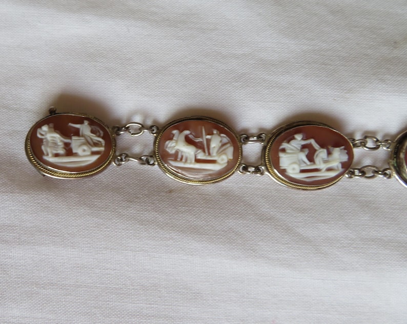 May include: A vintage bracelet featuring oval cameos with detailed white relief carvings set in gold-toned frames. The cameos depict classical figures and scenes, connected by delicate silver-toned chain links.