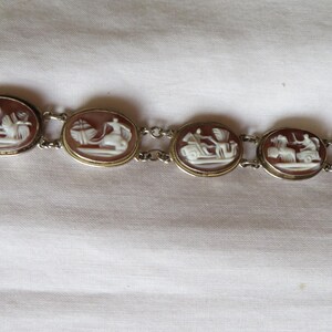 May include: A vintage bracelet featuring oval cameo pendants with white relief figures set in gold-toned frames. The bracelet is connected by delicate silver-toned chains and has a clasp closure. The cameos depict classical scenes.