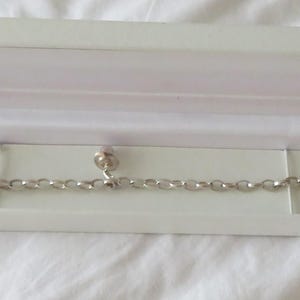 Könnte beinhalten: Ein silbernes Kettenarmband mit Anhänger, präsentiert in einer offenen weißen Geschenkbox. Das Armband hat ovale Glieder und einen kleinen Anhänger. Die rechteckige Box hat einen Klappdeckel.