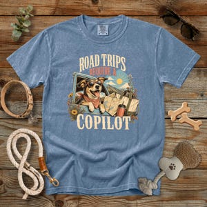 May include: A blue short-sleeve t-shirt featuring a graphic of a dog wearing sunglasses and a bandana inside a vehicle. The text reads ROAD TRIPS REQUIRE A COPILOT. The design includes a map, a travel mug, and sunflowers on a mountain background.