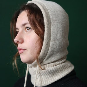 Può includere: Primo piano di una donna che indossa un passamontagna di lana beige con un lungo collo. Il passamontagna è realizzato in un materiale morbido e caldo e ha un design semplice.