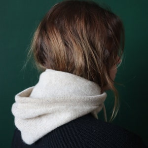 May include: A person with brown hair wearing a white hooded sweater over a black sweater.