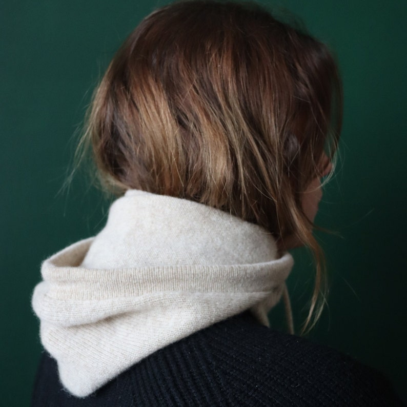 May include: A person with brown hair wearing a white hooded sweater over a black sweater.