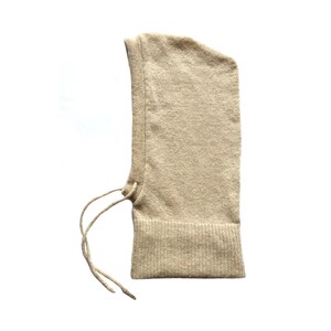 May include: A beige knitted balaclava with a drawstring tie. The balaclava is made of a soft, warm material and is ideal for cold weather.