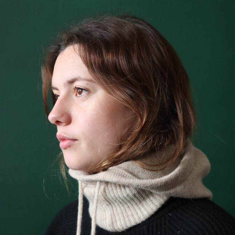 May include: A woman with brown hair wearing a beige knitted cowl neck sweater with a black shirt underneath. The woman is looking to the side.