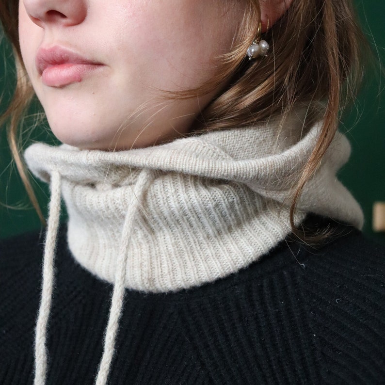 May include: A beige knitted cowl neck with a drawstring. The cowl neck is worn over a black sweater.