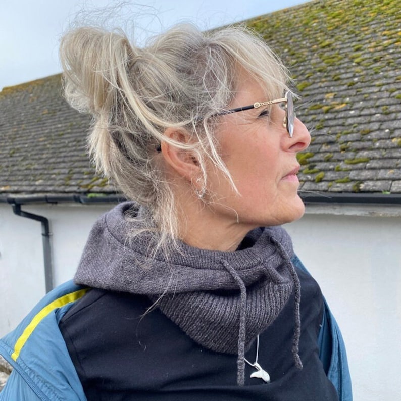 May include: A woman with grey hair wearing a grey hooded sweater, a black shirt, and a silver necklace with a whale tail pendant. She is wearing gold-rimmed glasses and looking to the right.