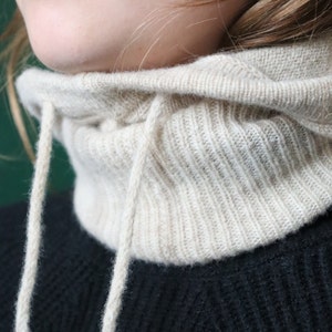 May include: A beige knitted cowl neck with a drawstring closure. The cowl neck is made of a soft, ribbed knit fabric.