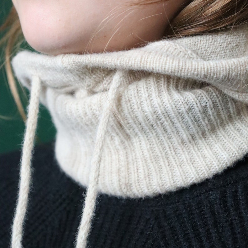 May include: A beige knitted cowl neck with a drawstring closure. The cowl neck is made of a soft, ribbed knit fabric.