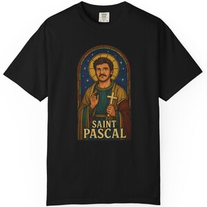 May include: Black t-shirt featuring a stained-glass style graphic of a man with a halo, holding a cross. The text "SAINT PASCAL" is below the image. The design is in shades of brown, gold, blue, and green.