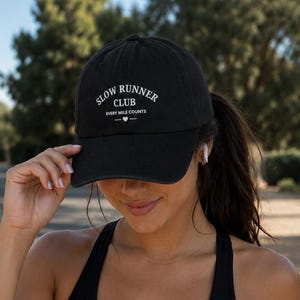 May include: Black baseball cap with the text "SLOW RUNNER CLUB" in white, with the words "EVERY MILE COUNTS" below. The cap is worn by a person wearing a black sports bra.