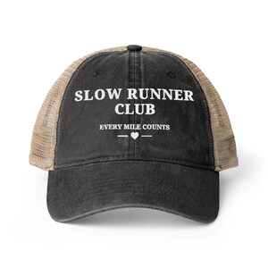 May include: A black trucker hat with a mesh back in a tan color. The front panel features white text that reads SLOW RUNNER CLUB, followed by the text EVERY MILE COUNTS with a small heart symbol centered between two horizontal lines below the text.