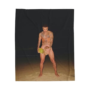 May include: A person standing on a beach holding two coconuts. The person has tattoos on their arms and chest. The background is dark, suggesting nighttime.