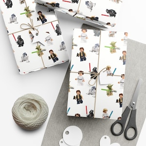 Pode incluir: Papel de embrulho branco com um padrão repetido de personagens Lego Star Wars, incluindo Darth Vader, Yoda, Luke Skywalker e Han Solo. O papel está embrulhado em torno de uma caixa de presente e amarrado com um barbante.