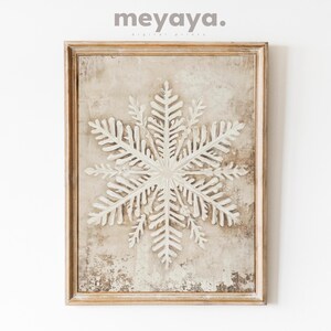 Puede incluir: Una impresión de arte de pared enmarcada con un copo de nieve blanco sobre un fondo beige. El marco es de madera clara con un acabado desgastado.
