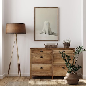 May include: A framed print of a white ghost with two black eyes on a light beige background. The print is hanging on a white wall above a wooden dresser with four drawers. A brown lamp with a tripod stand is on the left side of the dresser. A small potted plant is on the right side of the dresser.