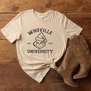May include: A beige t-shirt with a black graphic of the Grinch wearing a Santa hat. The graphic says "Whoville University" with "Est 1957" underneath.