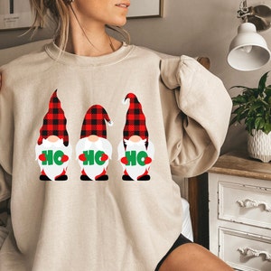 Könnte beinhalten: Ein beiger Pullover mit einem Aufdruck von drei Gnomen, die rote und schwarze karierten Hüte tragen und Schilder halten, auf denen "HO HO HO" steht.