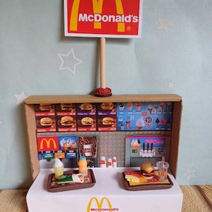 May include: A miniature cardboard replica of a McDonald's restaurant with a red and yellow sign that reads "McDonald's". The restaurant has a counter with a cash register, a drink dispenser, and a display of food items. There are two trays with miniature food items, including a burger, fries, and a drink.