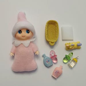 May include: A small doll wearing a pink fuzzy outfit and a matching pointed hat with a white collar. Accessories include a yellow woven basket, a white rectangular block, a yellow unicorn, two pacifiers, two small bottles, and a pink bottle.