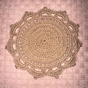 May include: A handmade, beige crochet doily with a circular design. The doily features a detailed, openwork pattern with a scalloped edge. The texture of the yarn is visible, adding to the handcrafted aesthetic. This decorative piece is suitable for home decor.
